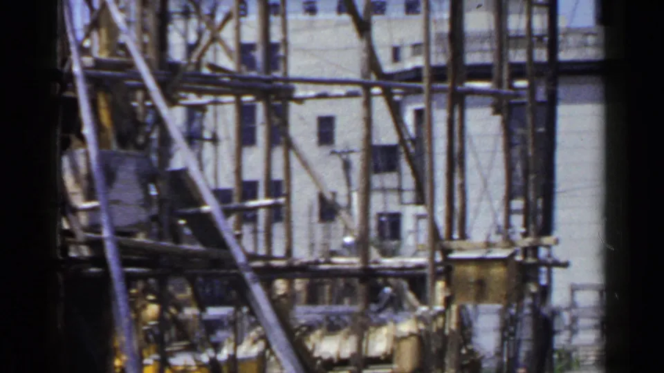 1953 Japan home-movie frame with dense wooden scaffold poles crossing a pale exterior wall with small windows.
