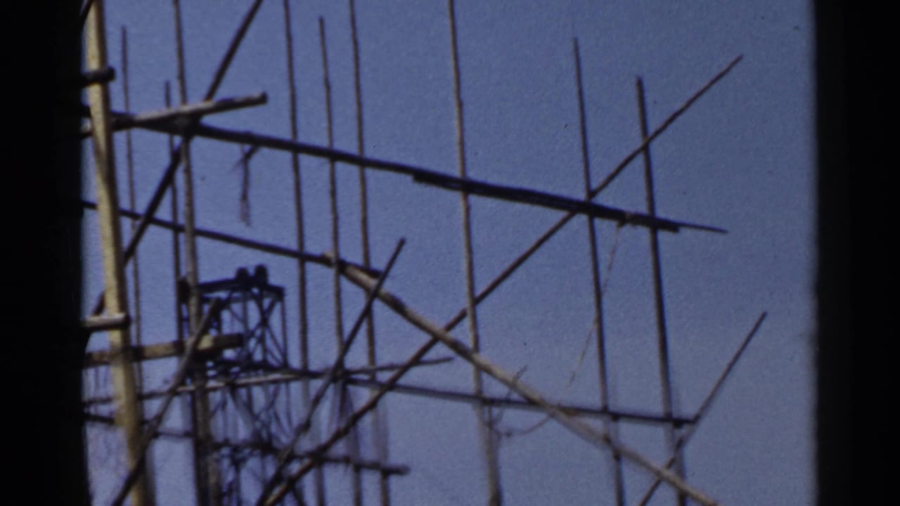 1953 Japan home-movie frame showing tall pole scaffolding, cross-bracing, and a distant worker on the structure.