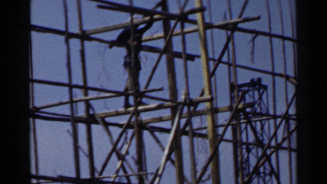 1953 Japan frame showing a construction worker positioned high within a dense bamboo-like scaffolding structure.