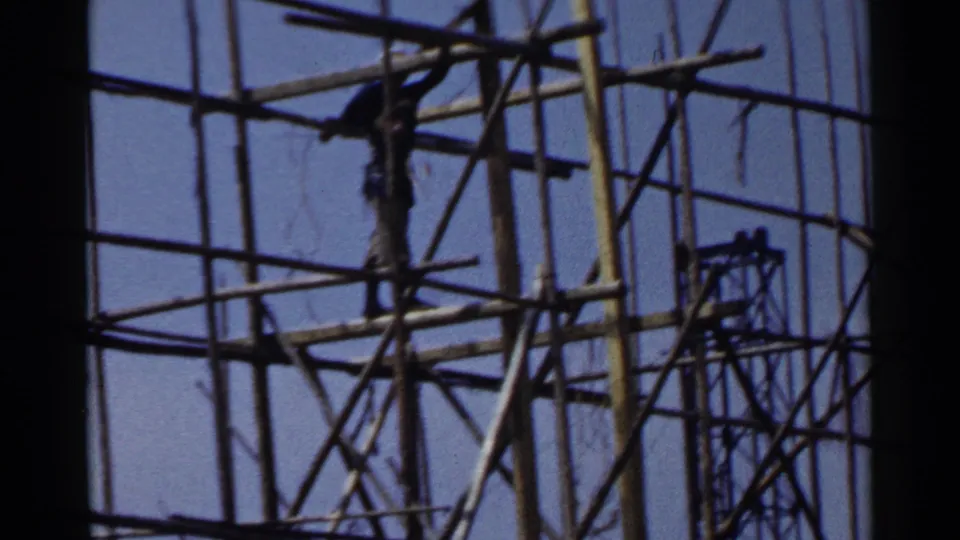 1953 Japan frame showing a construction worker positioned high within a dense bamboo-like scaffolding structure.