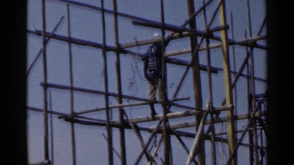 1953 Japan 8mm frame showing a worker in work clothes standing on a scaffold platform within a dense construction frame.