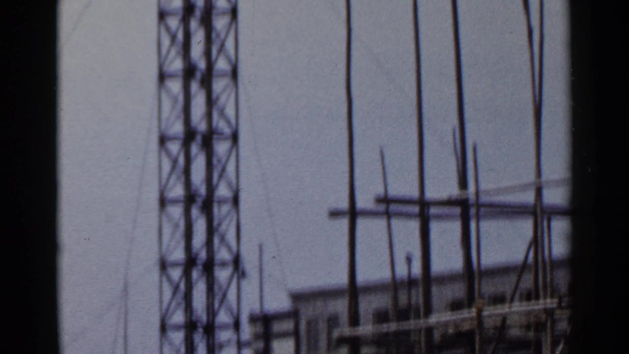 1953 Japan 8mm frame showing a lattice radio tower, tall poles, scaffolding, and part of a work-site building.