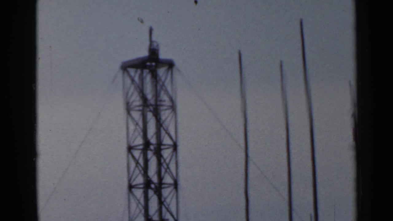 A 1953 Japan home-movie frame centers a lattice radio tower with several vertical poles in the foreground.