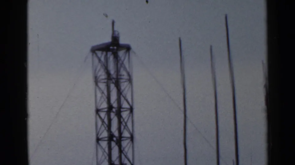 A 1953 Japan home-movie frame centers a lattice radio tower with several vertical poles in the foreground.