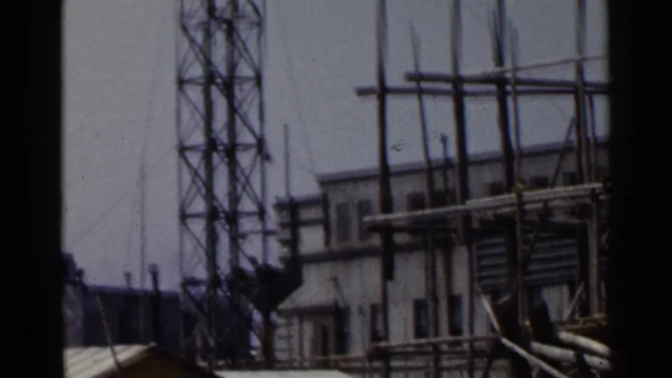 1953 Japan construction-site frame with a worker near scaffolding, a lattice tower, poles, and a low building.