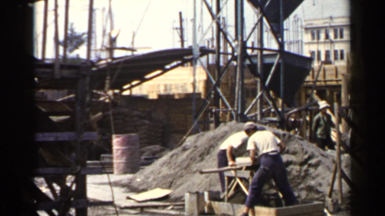 In Japan 1953 footage, adult workers in light shirts handle materials near scaffolding, wooden forms, a red barrel, and a mound of sand or aggregate.