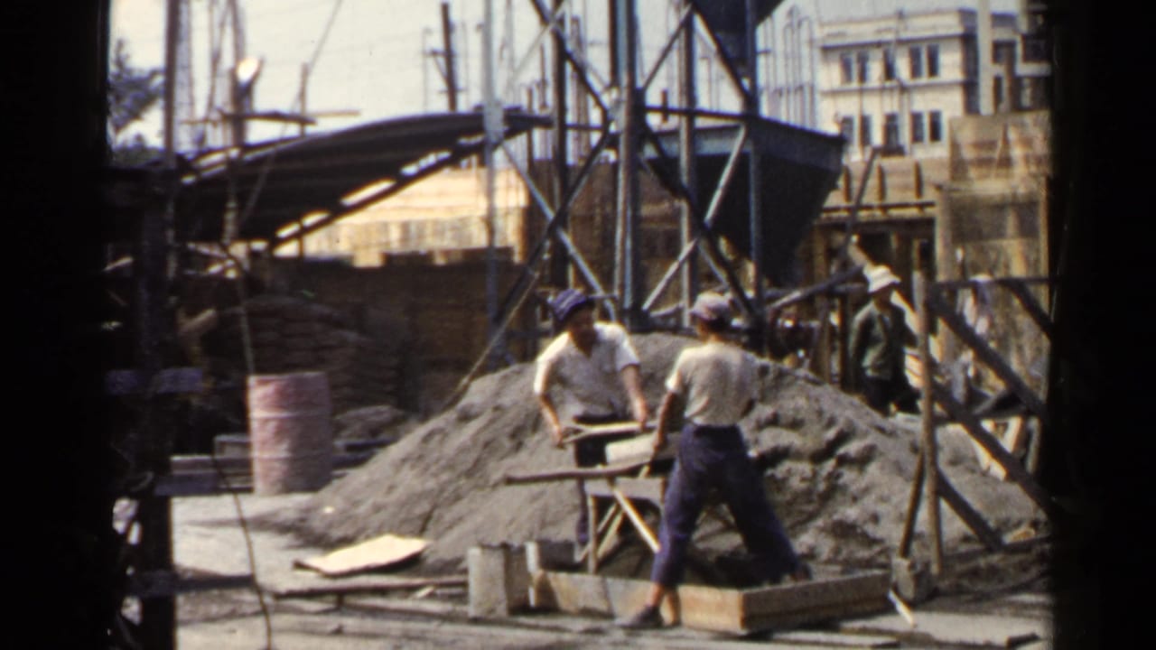 Two workers stand near a mixing table while scaffolding and building facades fill the background.