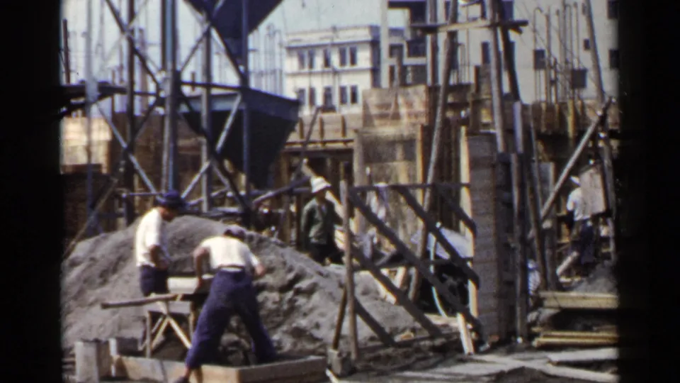 The final frame shows the Japan construction site with workers around a mixing area, timber supports, scaffolding, scattered planks, and urban buildings beyond.