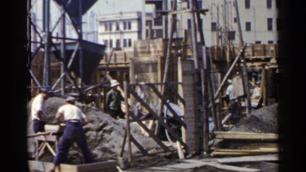 Workers stand and move around a construction site with timber supports, gravel, and nearby buildings.