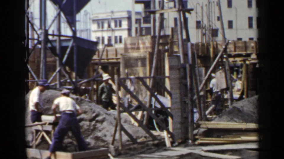 1953 Japan home-movie frame of men working at an urban construction site with wood formwork, supports, gravel, and background buildings.