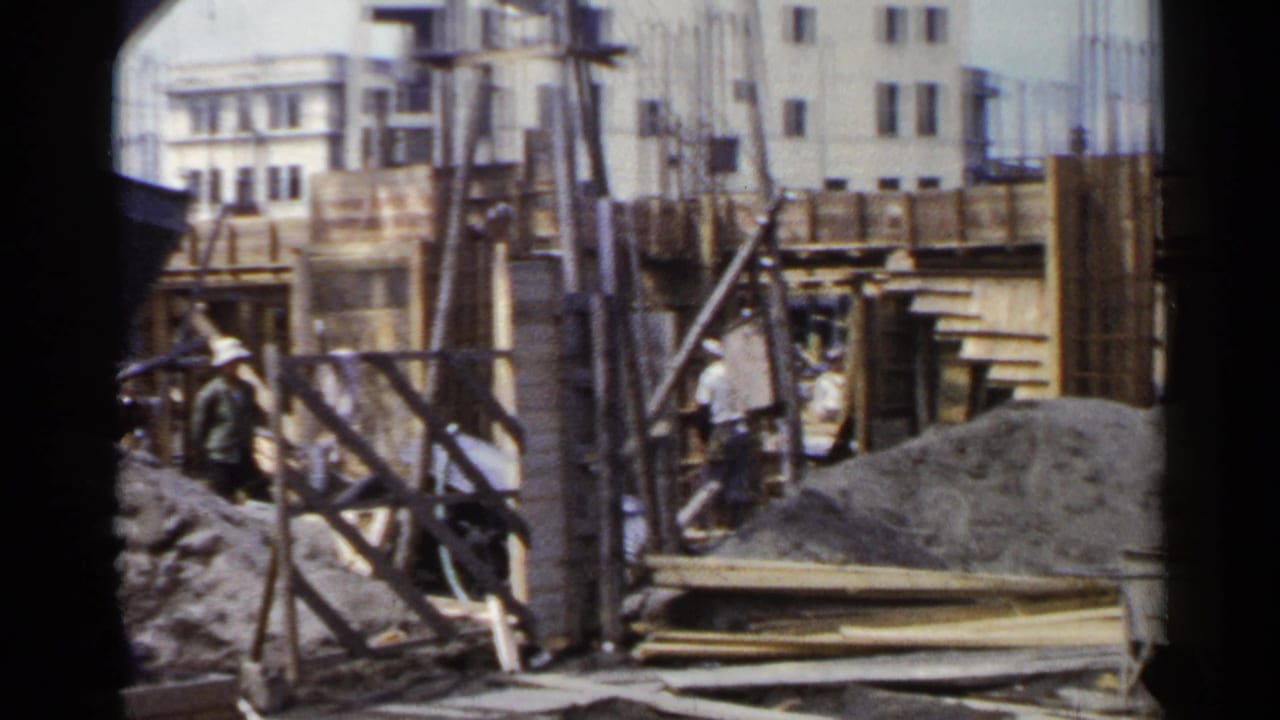 1953 Japan frame showing workers inside a building site with exposed rebar, timber formwork, gravel, and urban buildings behind.