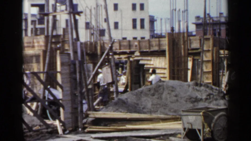 1953 Japan construction-site frame with workers partly obscured by formwork, vertical rebar, planks, gravel, and a building facade in back.