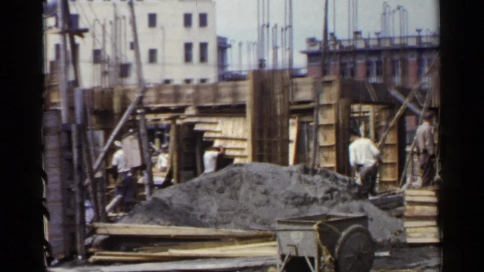 1953 Japan home-movie frame of men at a construction site with wood forms, rebar, planks, gravel, and a wheelbarrow in the foreground.