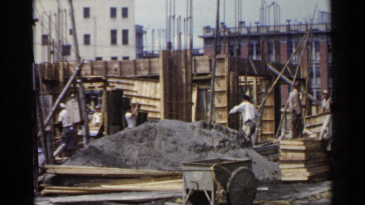 Workers move around wood formwork, rebar, gravel, and a wheelbarrow at an urban construction site.