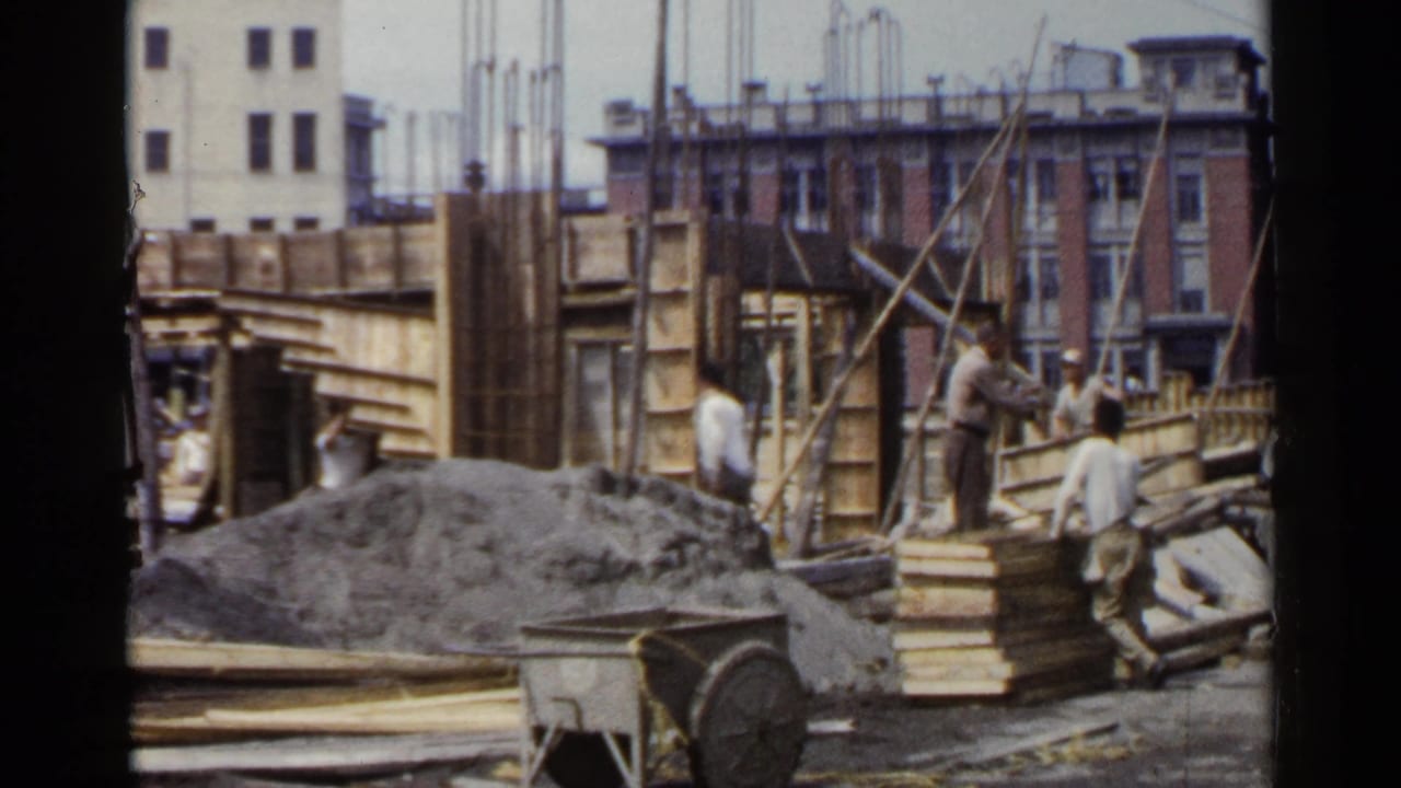 Men work beside timber framing and rebar with gravel, boards, and city buildings in the background.