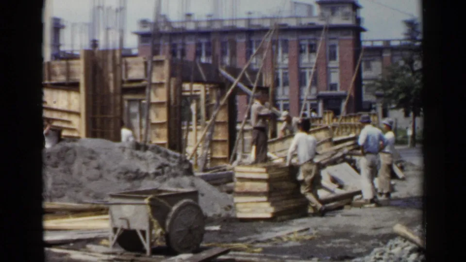 1953 Japan home-movie footage shows workers beside timber formwork, vertical rebar, lumber stacks, gravel, and a wheelbarrow.
