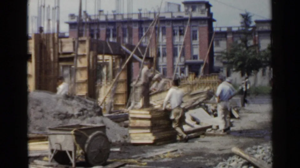 At a 1953 Japan construction site, workers appear among wooden formwork, exposed rebar, gravel, and urban buildings.