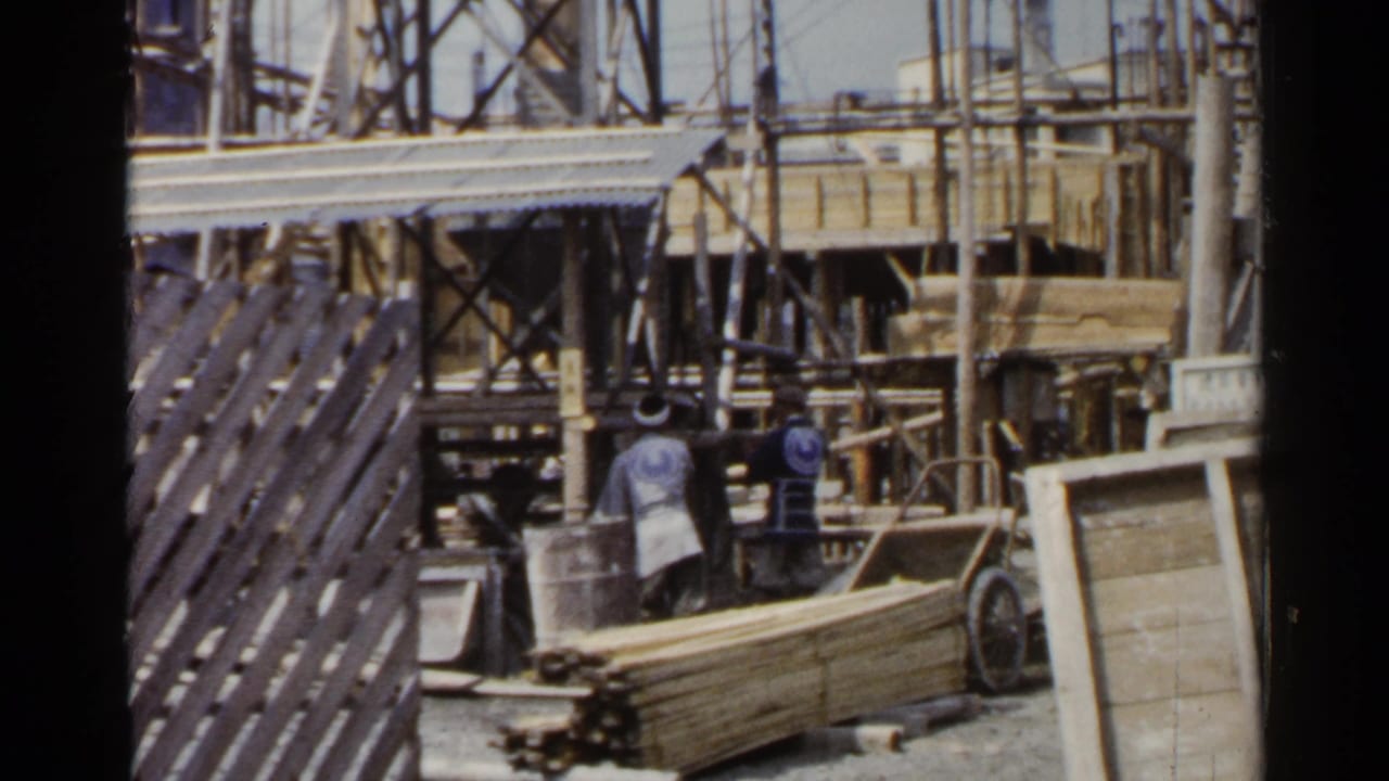Workers stand among scaffolding, stacked lumber, and a handcart at a construction site.