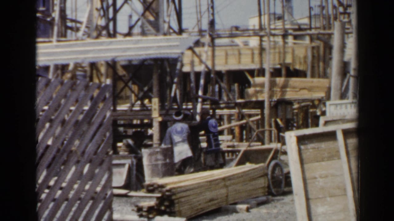 1953 Japan home-movie frame with workers beside stacked boards, scaffolding, and partially built structures.