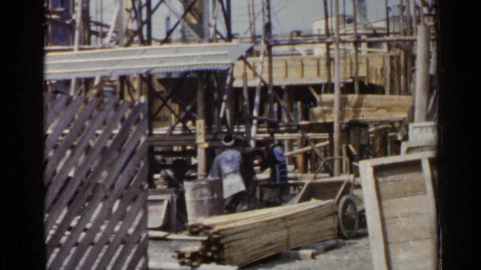 1953 Japan 8mm view of workers near a handcart, lumber stacks, and layered construction scaffolding.