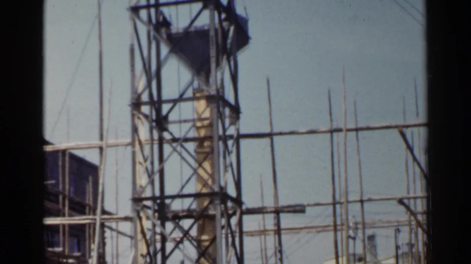 1953 Japan home-movie frame with tall scaffolding, vertical poles, partial building edges, and open sky.