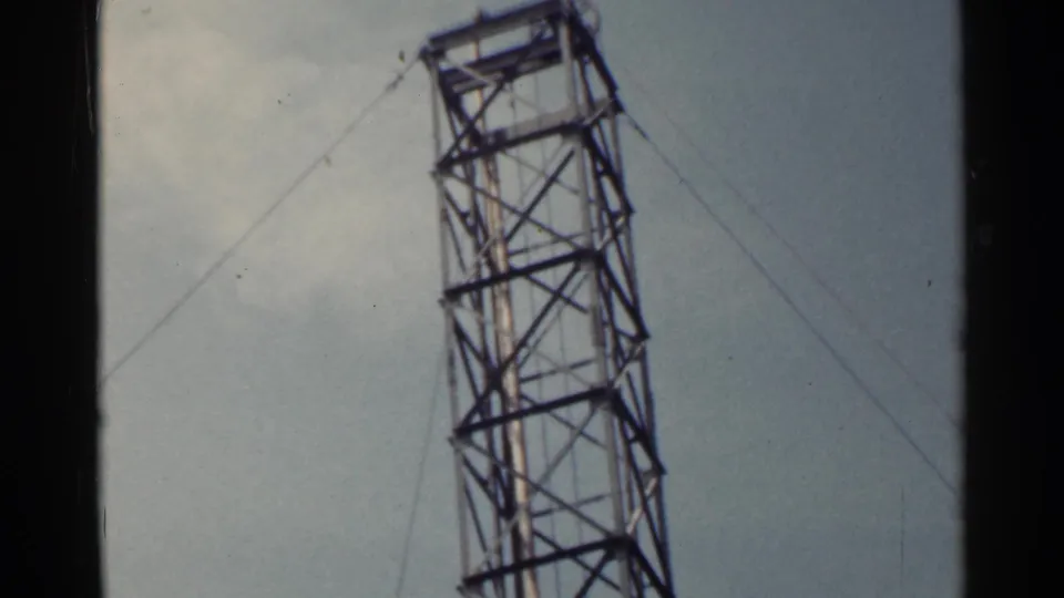 1953 Japan home-movie frame with an angled lattice tower and support cables above a building site.