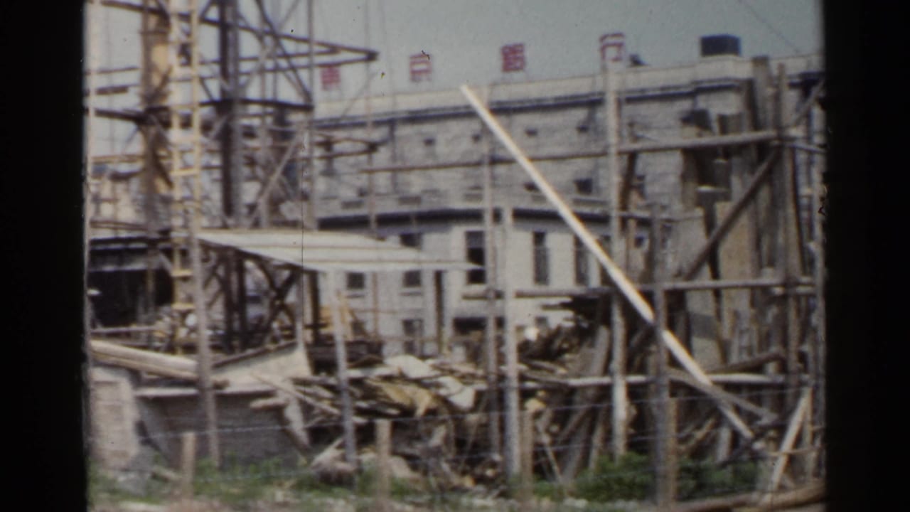 1953 Japan frame showing a construction area with timber bracing, stacked materials, and a large building behind it.