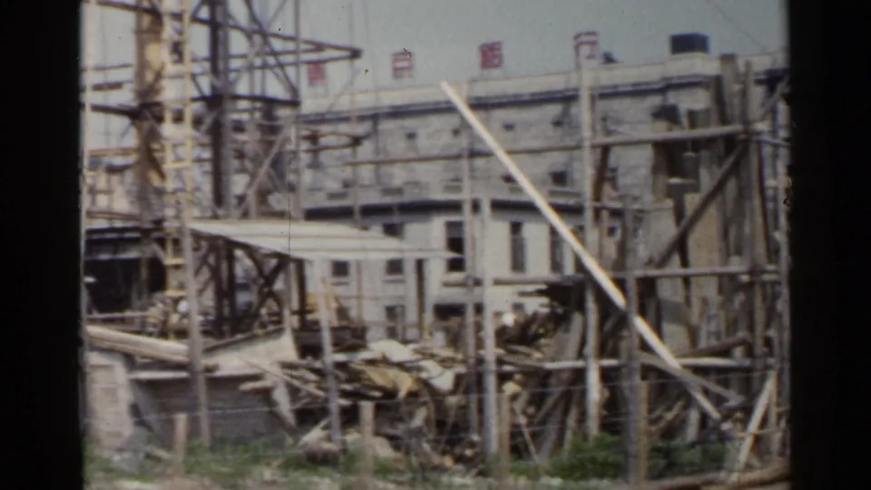 1953 Japan frame showing a construction area with timber bracing, stacked materials, and a large building behind it.