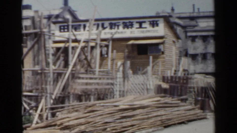 1953 Japan 8mm frame showing a sign-marked construction hut, timber scaffolding, and long poles stacked across the foreground.