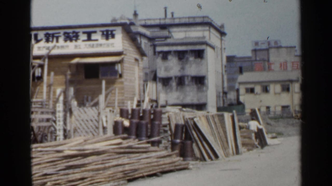 1953 Japan frame of a construction yard with timber piles, a sign in Japanese characters, scaffolding, and neighboring urban buildings.