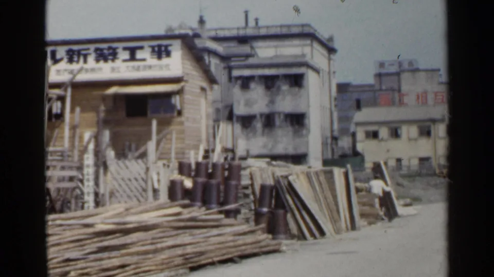 1953 Japan frame of a construction yard with timber piles, a sign in Japanese characters, scaffolding, and neighboring urban buildings.