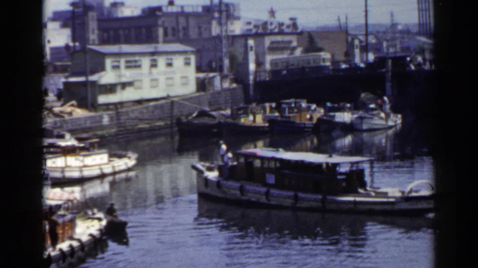 1953 Japan 8mm frame showing small boats on a river or canal with a bridge, waterfront buildings, and distant traffic.