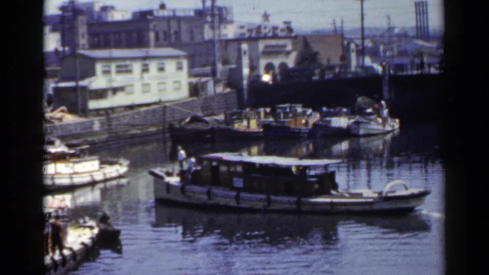 1953 Japan frame showing a boat passing on a river or canal with other craft, a bridge, industrial buildings, and faint people visible.