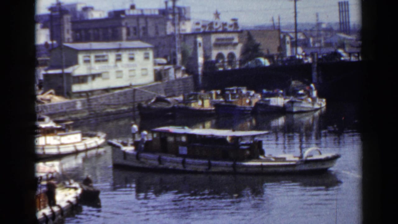 1953 Japan canal view with small boats, a larger houseboat-like vessel, industrial buildings, utility poles, and water reflections.