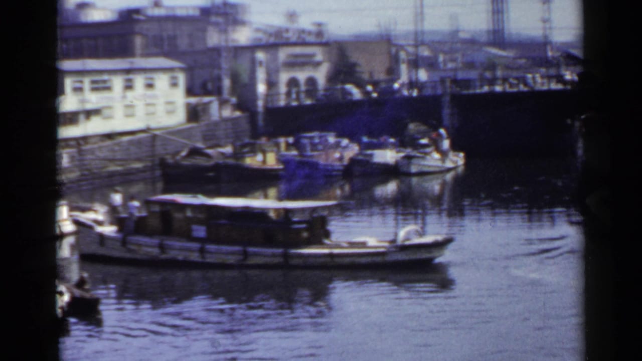 A low passenger or houseboat-like craft moves across a Japanese city canal, with docked boats, buildings, and poles behind it.