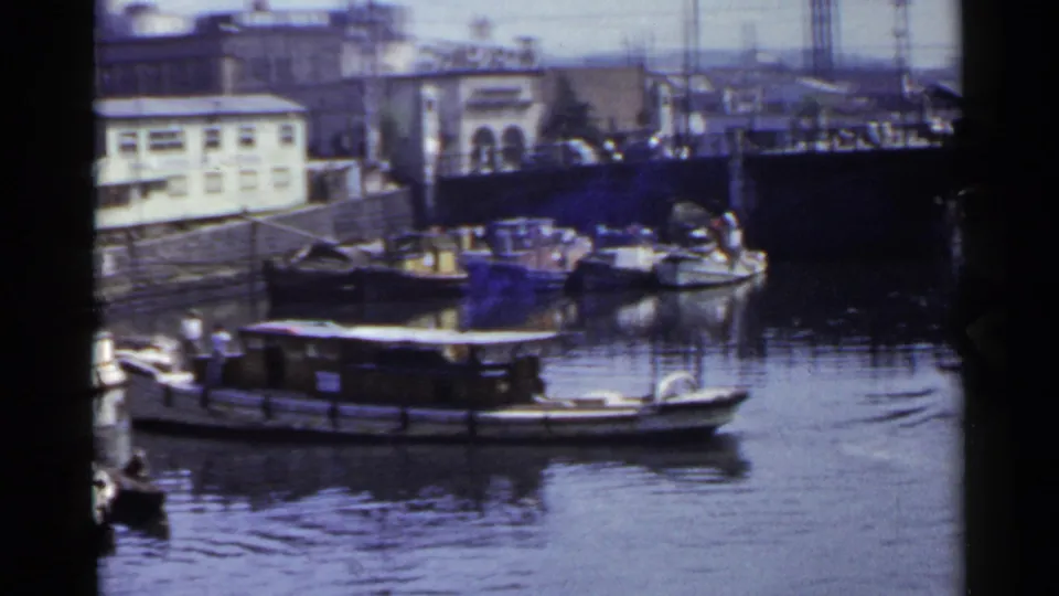 A low passenger or houseboat-like craft moves across a Japanese city canal, with docked boats, buildings, and poles behind it.