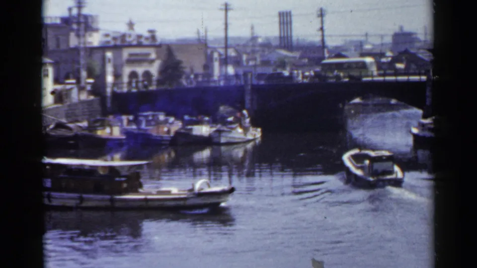 A small motorboat moves near an arched bridge on a 1953 Japanese city canal, with larger boats and waterfront buildings nearby.