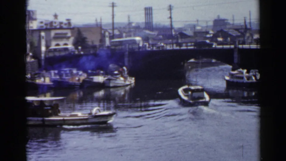 A small motorboat travels away through a city canal in Japan, passing under or near an arched bridge with docked boats alongside.