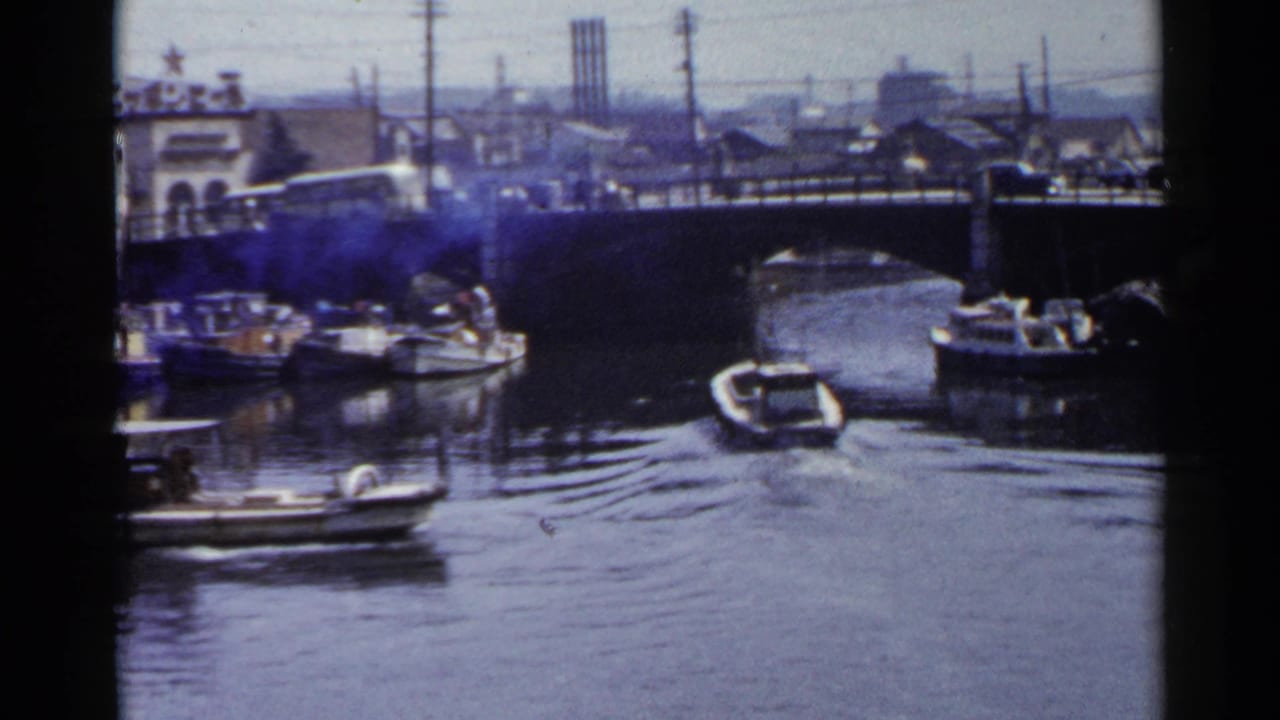 1953 Japan 8mm frame showing motorboats on a canal near a bridge with vehicles, utility poles, buildings, and distant smokestacks.