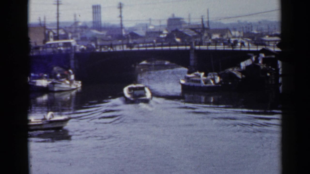 1953 Japan 8mm frame with a boat moving up a canal toward a bridge, with buses or cars, poles, buildings, and industrial stacks in the background.