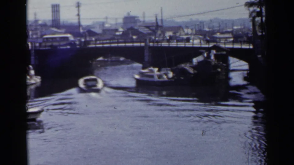 1953 Japan 8mm frame showing canal boats near a bridge lined with traffic, with urban buildings, utility poles, and distant smokestacks visible.