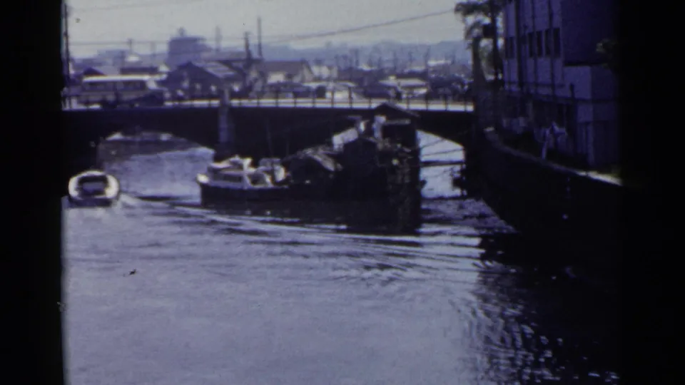 1953 Japan 8mm frame with a small canal boat near a bridge, road traffic above, and a darker building edge framing the right side.