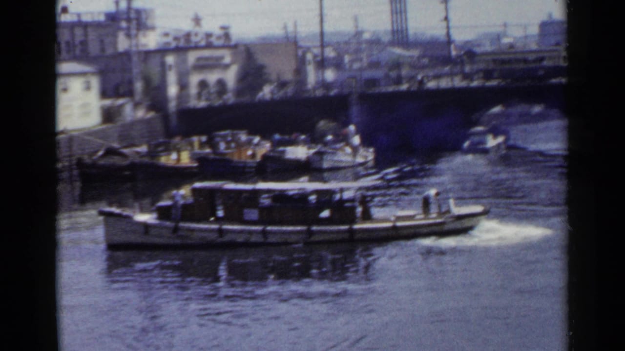 Small motorboat moving across a canal with docked boats, bridge structure, and waterfront buildings behind it.