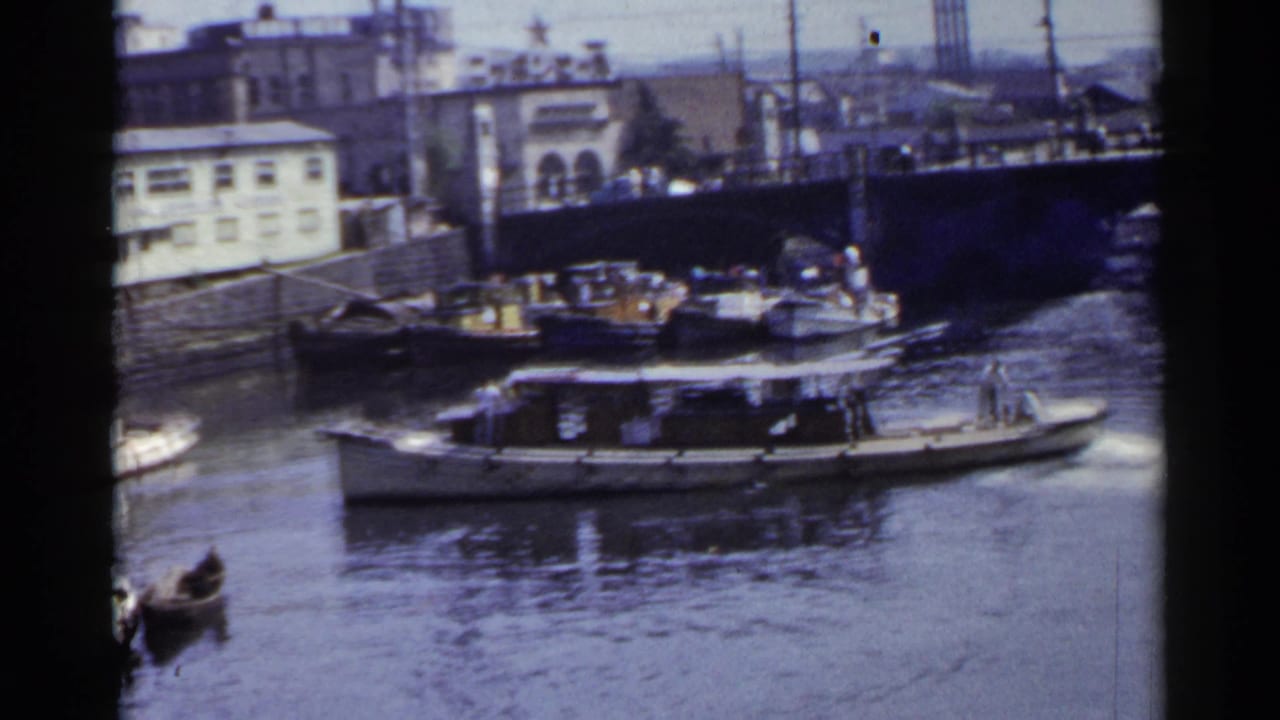 1953 Japan home-movie frame with a motorboat traveling past docked wooden vessels and a small rowboat on a canal.