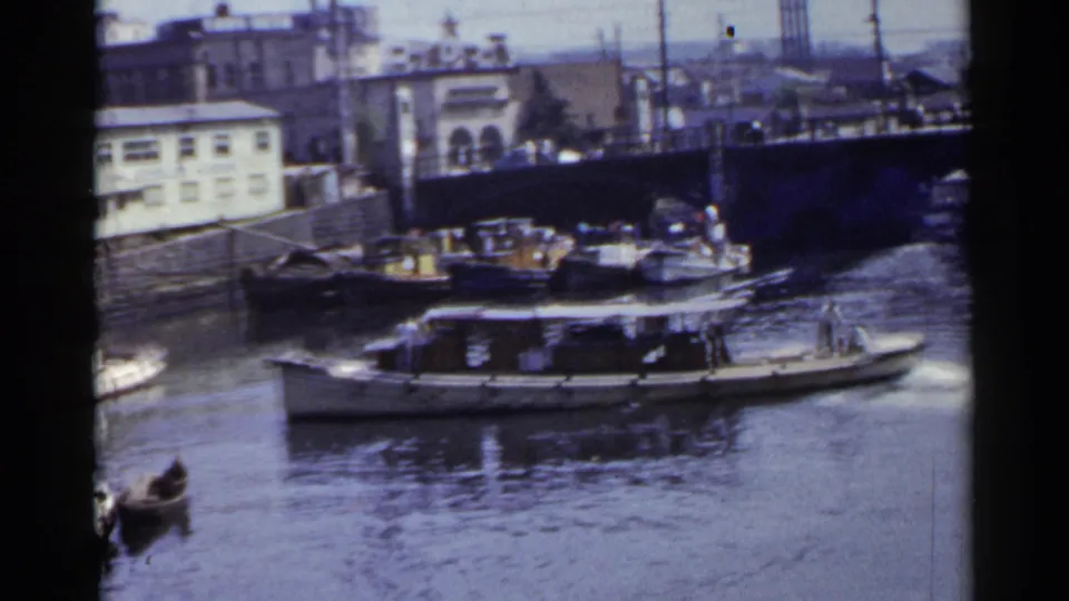 1953 Japan home-movie frame with a motorboat traveling past docked wooden vessels and a small rowboat on a canal.