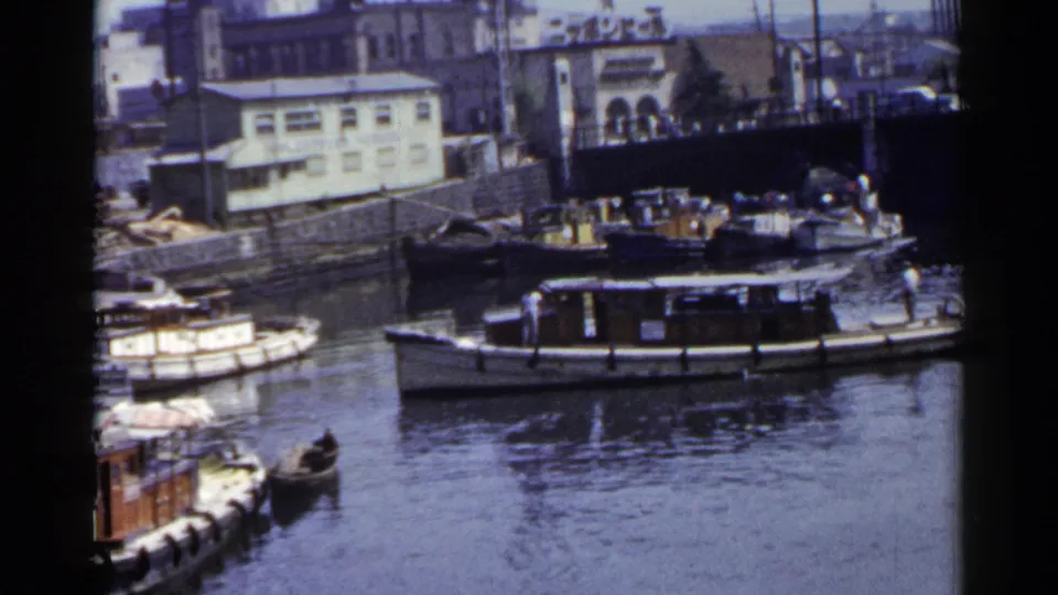 1953 Japan 8mm view of a passenger motorboat passing moored boats along a canal lined with buildings and stonework.