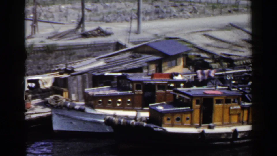1953 Japan home-movie frame showing moored wooden boats along a canal edge, with rooftops, portholes, and hanging cloth visible.