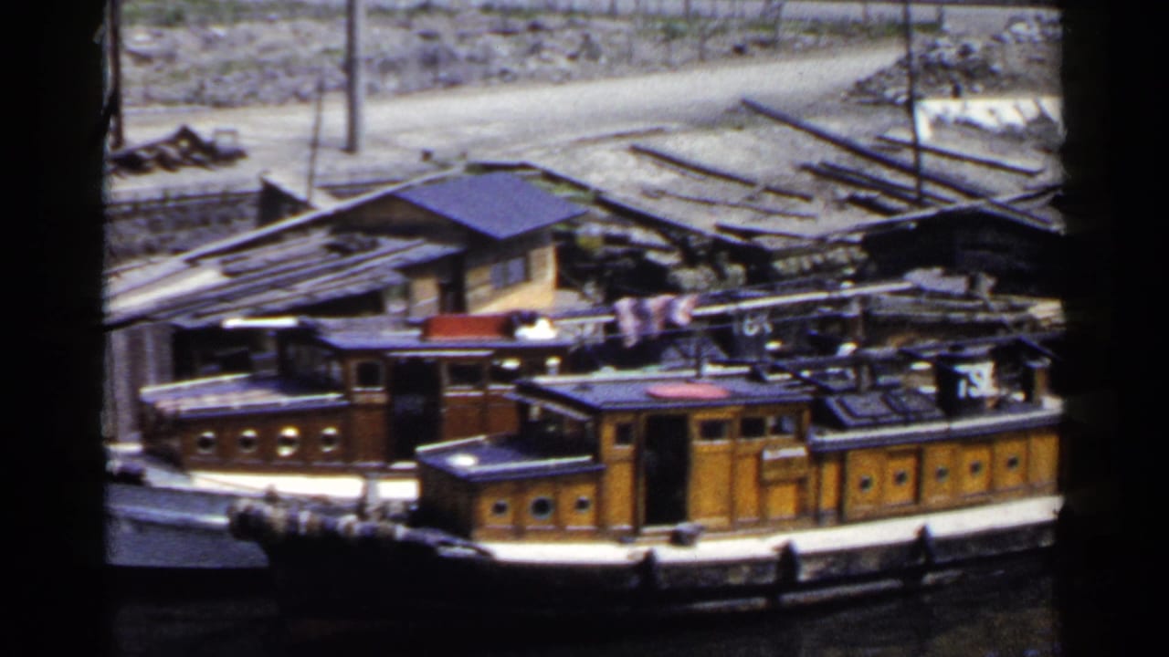 1953 Japan home-movie frame showing weathered wooden boats tied along a riverbank below rough sheds and scattered shoreline material.