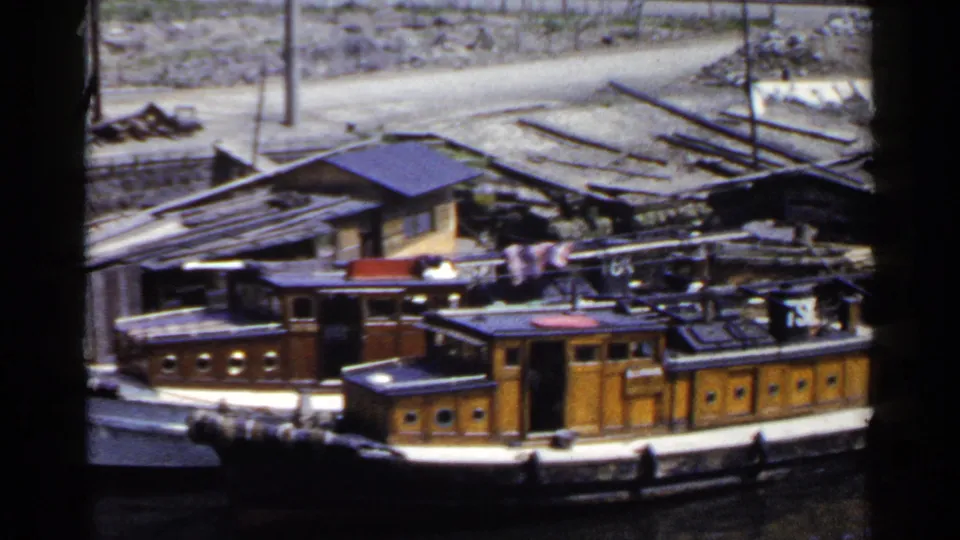 1953 Japan home-movie frame showing weathered wooden boats tied along a riverbank below rough sheds and scattered shoreline material.