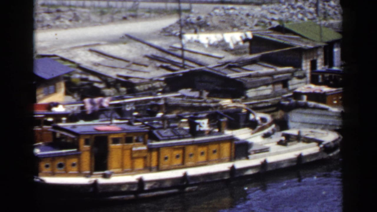 1953 Japan riverside view with functional wooden boats, dark water, rough sheds, and scattered material along the bank.